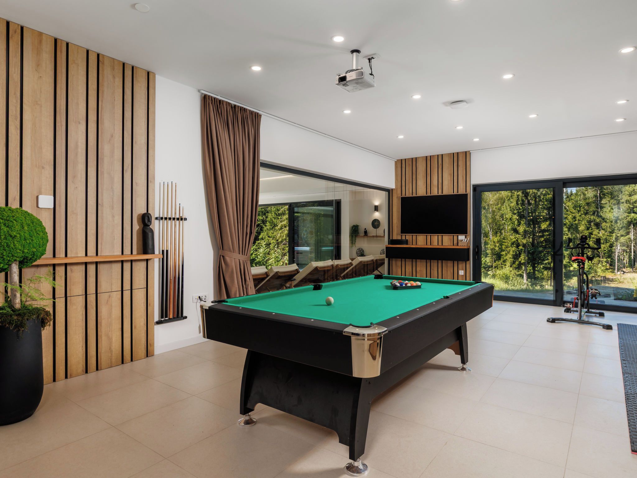 A modern indoor pool table with a green felt surface, surrounded by wooden paneling and a large window offering a view of a forested area.