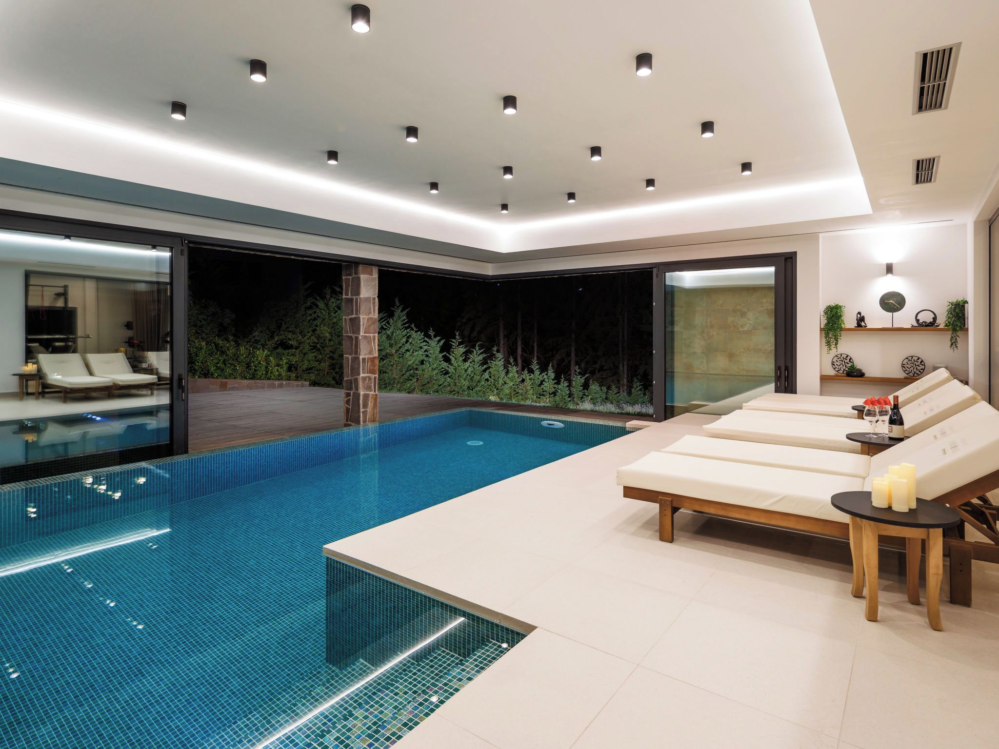 Una piscina interna moderna con zone relax, una parete in vetro e vista su una zona boschiva.