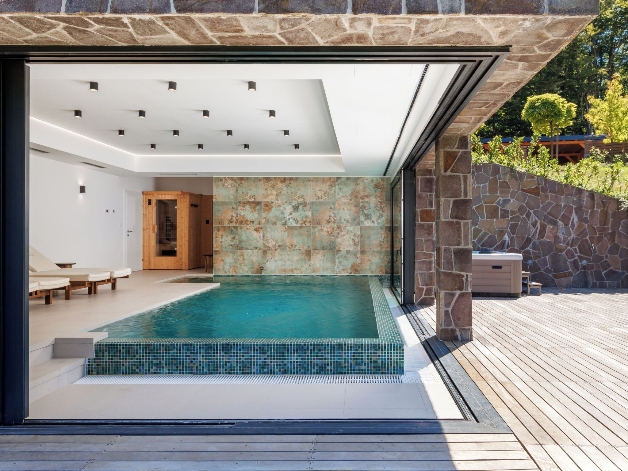 Une piscine intérieure moderne, avec un mur en pierre en arrière-plan, une terrasse en bois et un sauna.
