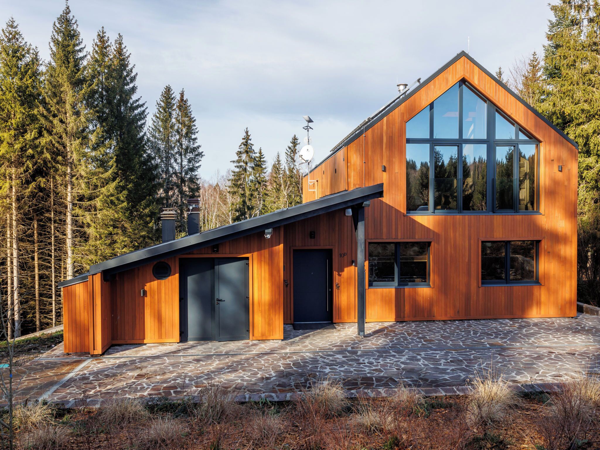 A modern wooden house with a large glass window, a stone-paved driveway, and a forested backdrop.