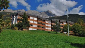 Um hotel com vários andares e telhado plano, rodeado por um gramado bem cuidado e árvores, com montanhas ao fundo.