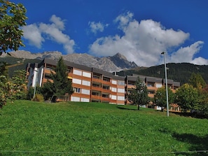 Ein mehrstöckiges Hotel mit flachem Dach, umgeben von einer gepflegten Rasenfläche und Bäumen, mit Bergen im Hintergrund.