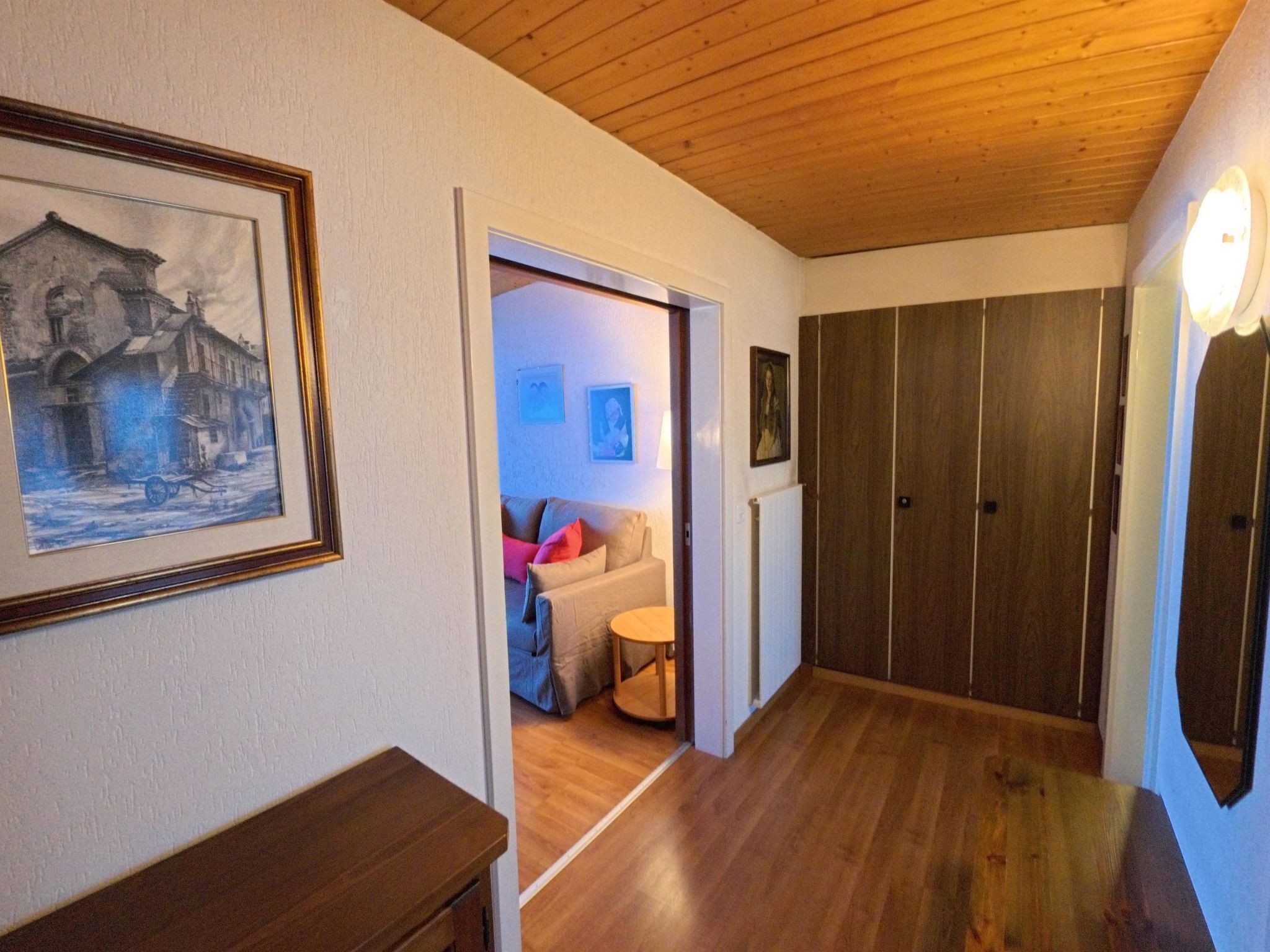A hallway with wooden flooring, a framed painting on the left, and a door leading to a room with a sofa and a small table.