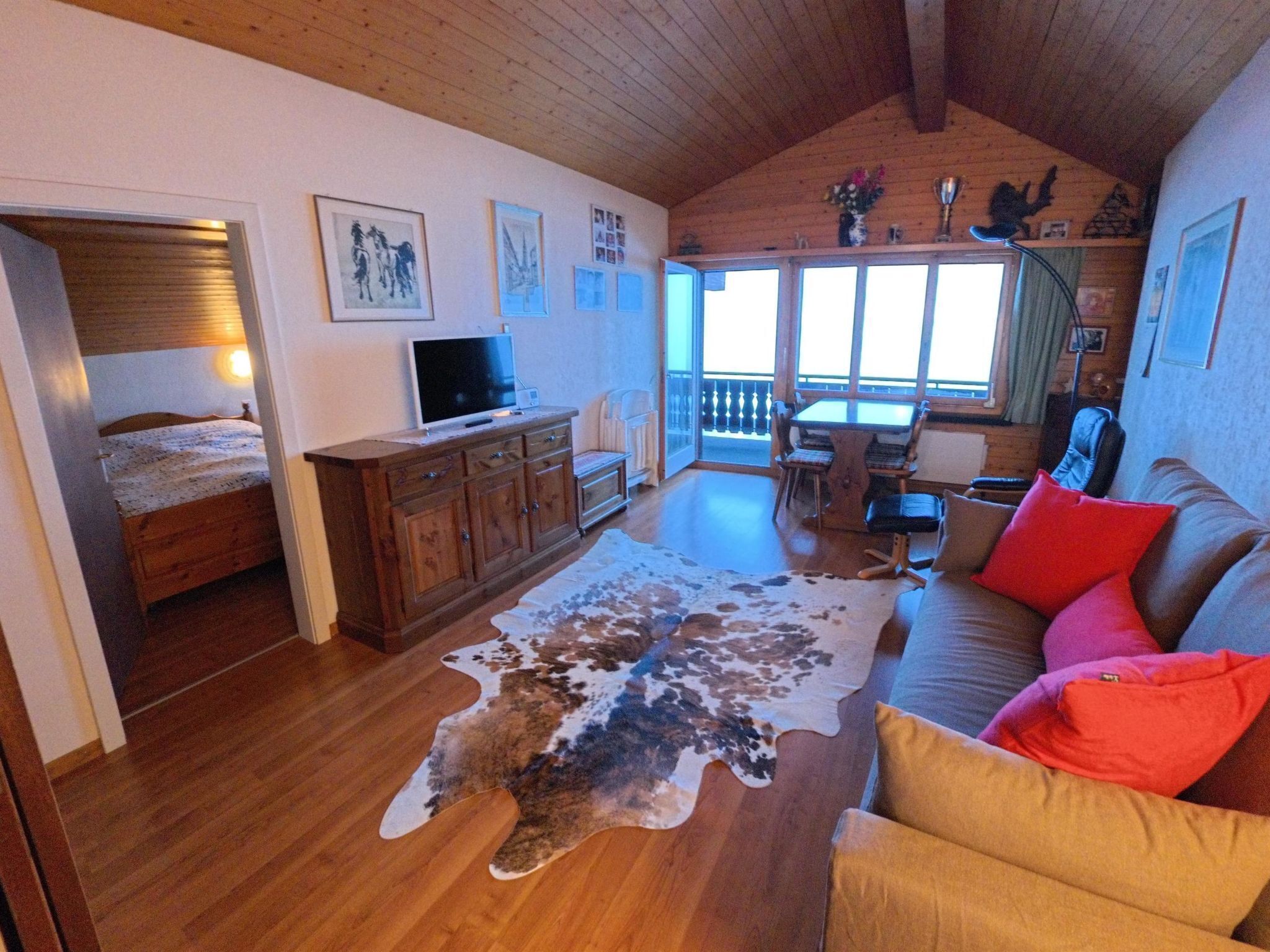 A cozy living room with a wooden ceiling, a large window, a cowhide rug, a sofa with colorful cushions, a TV, and a wooden cabinet.