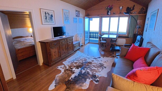 Ett mysigt vardagsrum med ett träpanelat tak, ett stort fönster, en matta av kohud, en soffa med färgglada kuddar, en tv och ett träskåp.