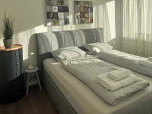 A modern bedroom with a grey upholstered headboard, two single beds with white linens, a small bedside table with a lamp, a dark dresser, and a potted plant. - Tgesa Tgampi T20 Appartement/Fewo, Dusche, WC, 2 Schlafräume by Interhome (Savognin)