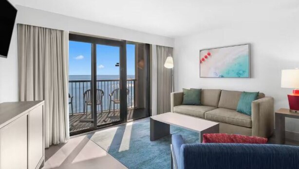 A hotel room with a balcony, a sofa, a coffee table, and a TV. - Compass Cove Beachfront Escape | Pool Access | Balcony | Hot Tub | Free Parking (Myrtle Beach)