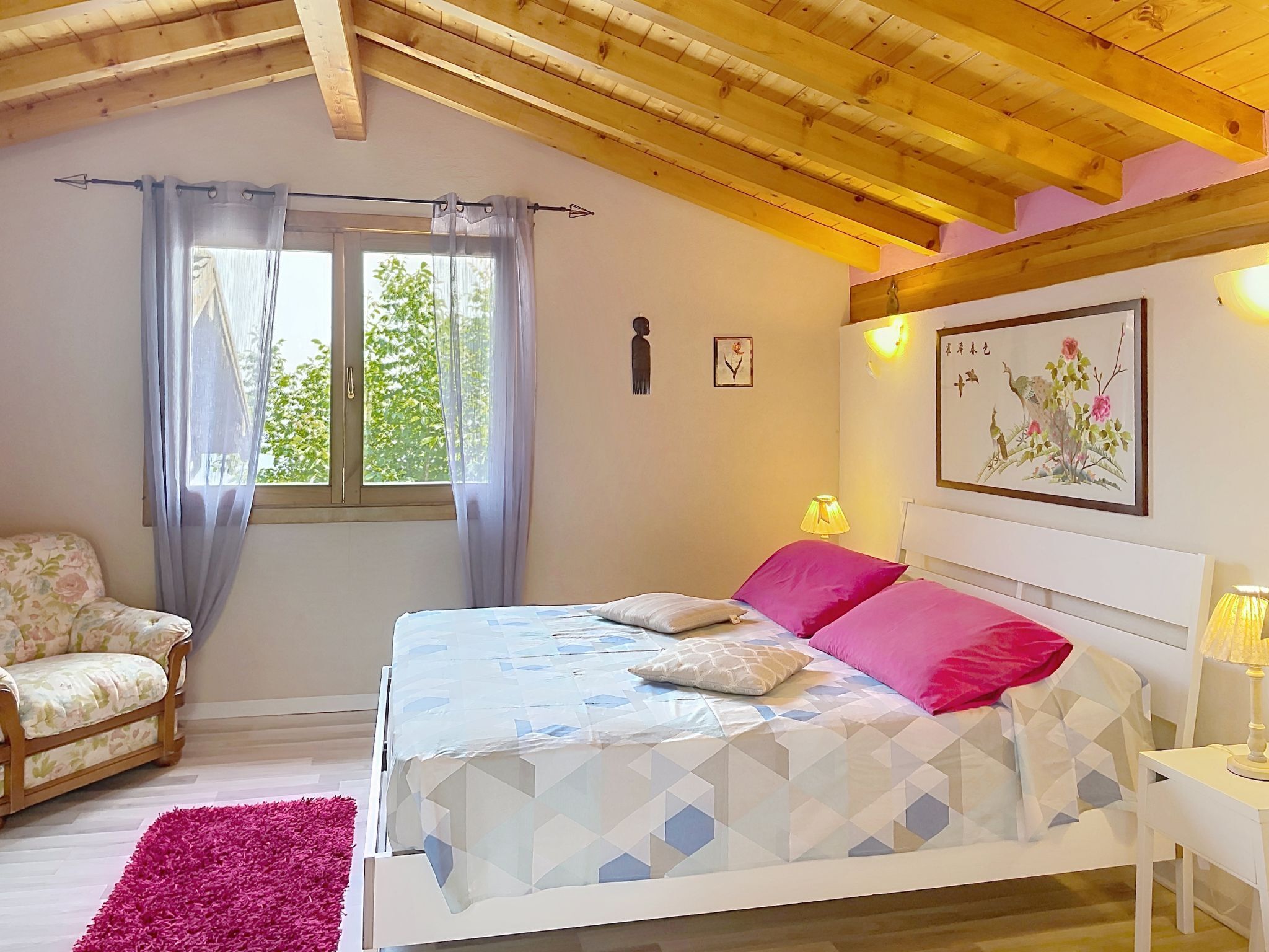 Un dormitorio con cama, una silla, ventana con cortinas, pared con un cuadro y techo de madera.