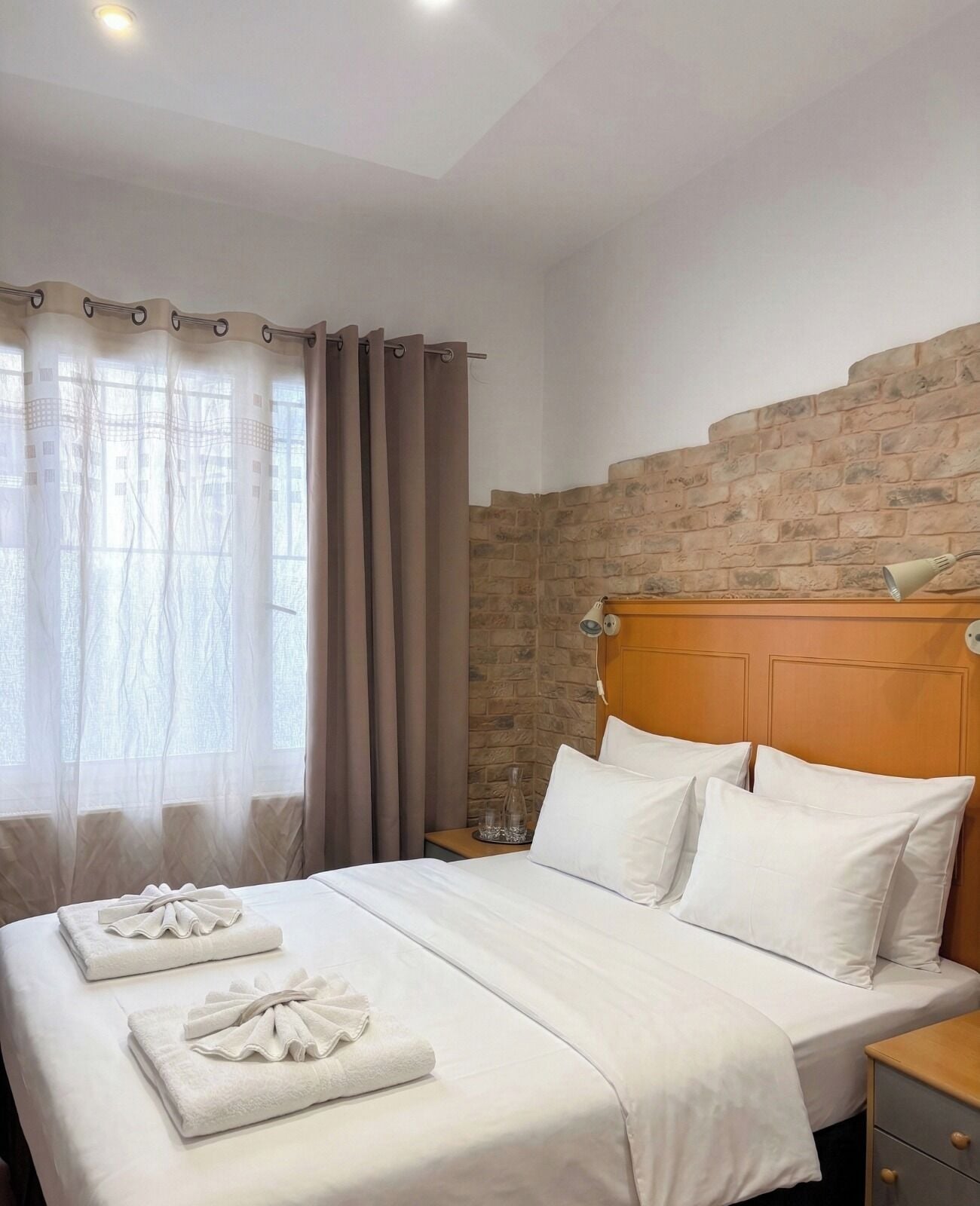 A hotel room with a neatly made bed, a headboard with a stone-like texture, and a window with sheer curtains.