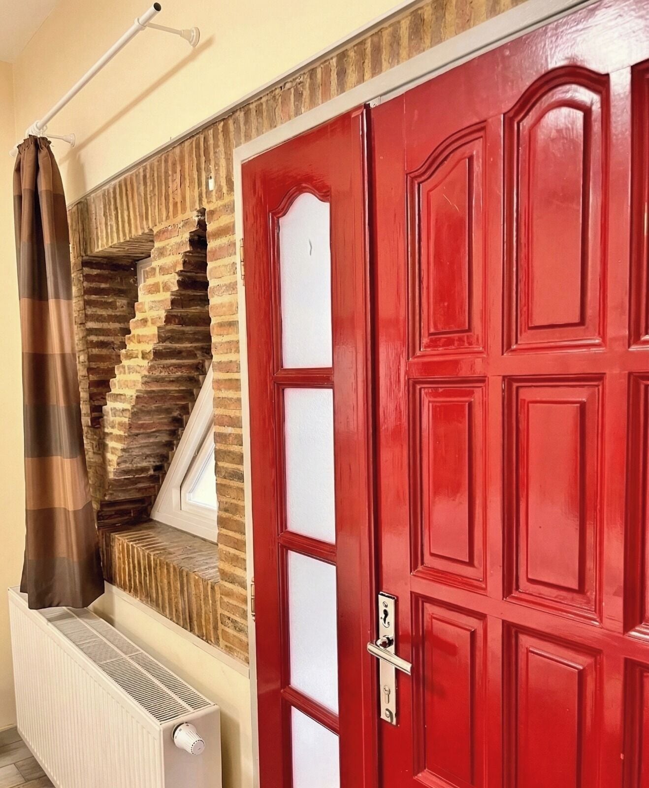 Una puerta roja con mirilla, un radiador blanco y una cortina marrón a rayas.