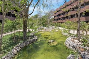 A landscaped area with a clear water feature surrounded by stone borders, trees, and a modern building with balconies. - Luxury Condo at Amira District Rooftop Infinity (Tulum Municipality)