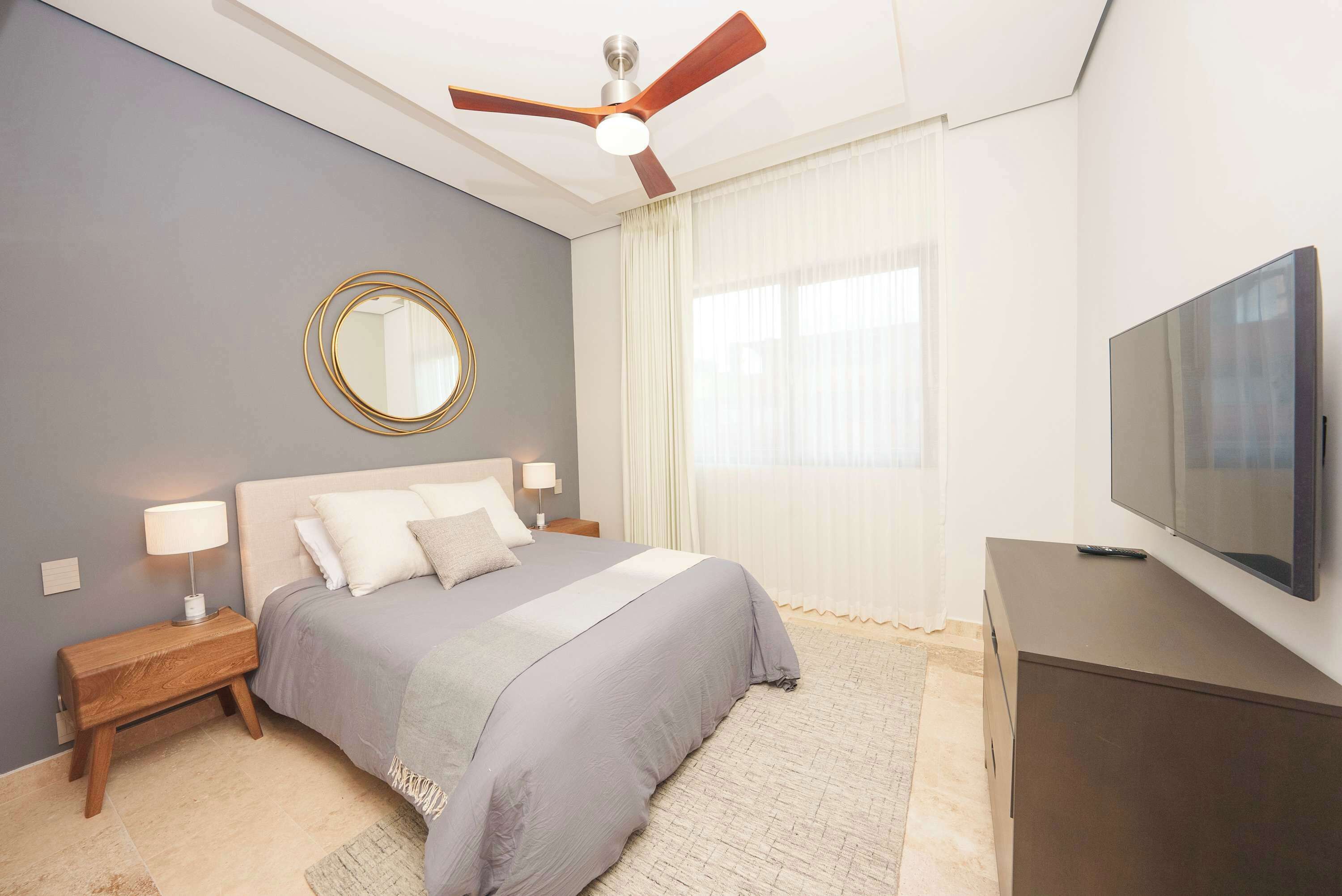 Une chambre à coucher comprenant un lit, des tables de chevet, un ventilateur de plafond, une télévision et une grande fenêtre avec des rideaux.