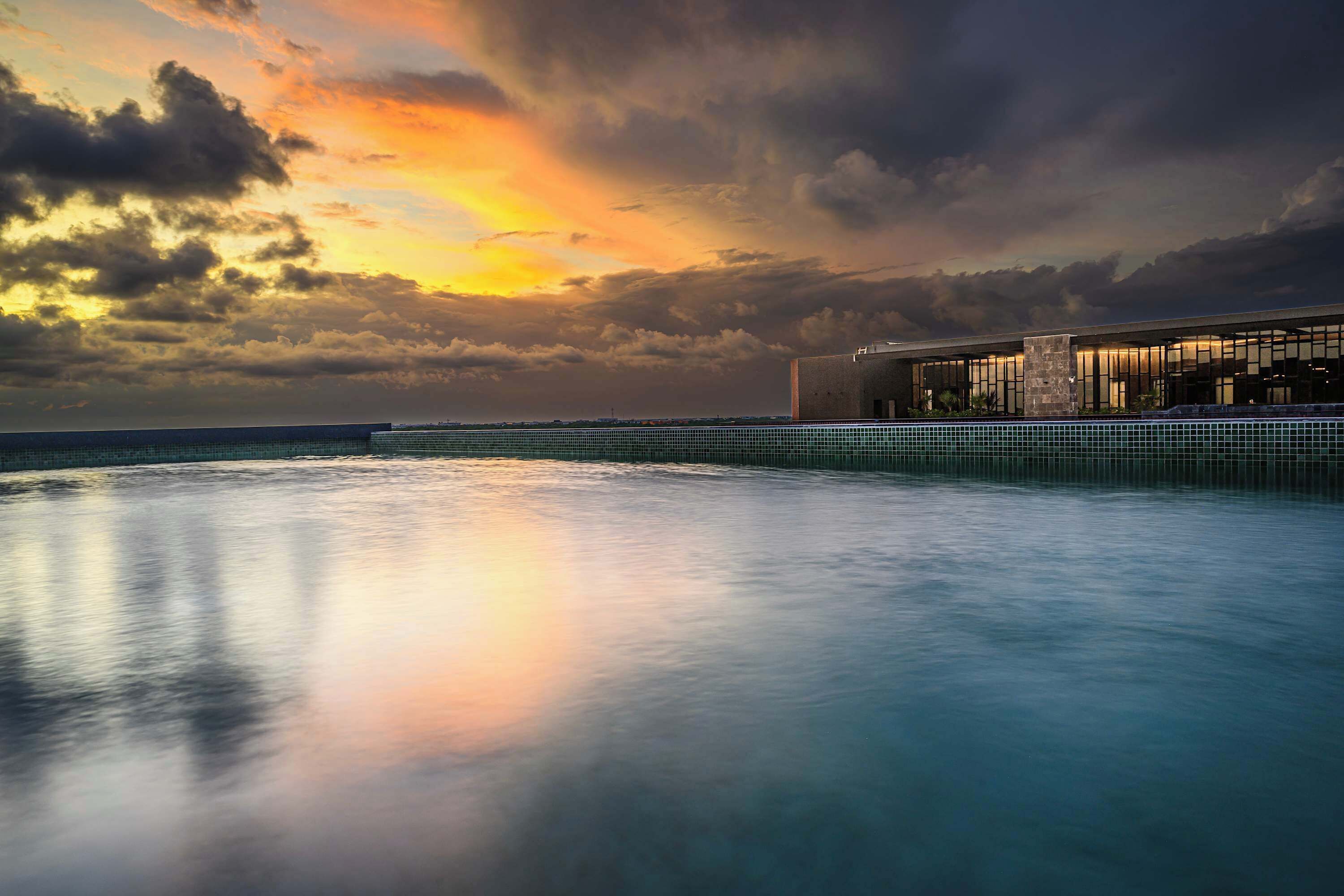 A modern building with a pool in the foreground and a sunset in the background.