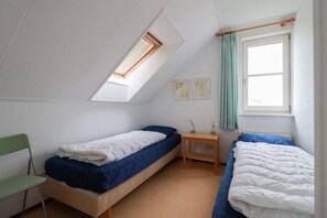 Ein kleines, kompaktes Zimmer mit zwei Einzelbetten, einem Tisch, einem Stuhl, einem Fenster mit Vorhängen und einem Oberlicht.