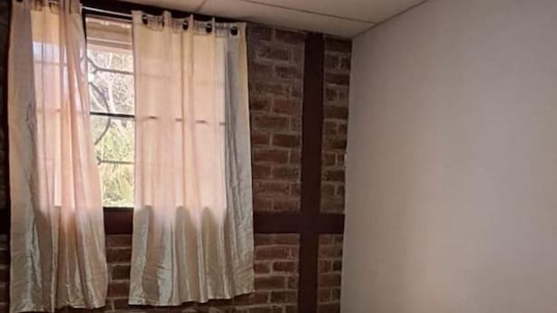 Une petite chambre avec un lit simple, un mur en briques, une fenêtre avec des rideaux et une petite table en bois.