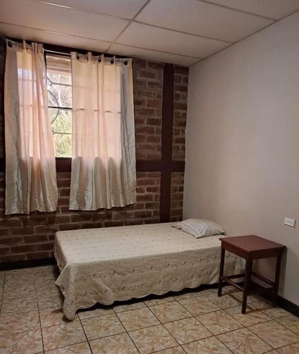 A small, single-bed room with a brick wall, a window with curtains, and a small wooden table.