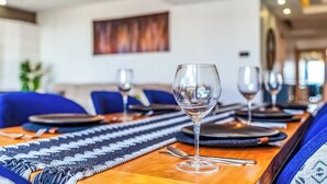 Una mesa de comedor dispuesta con copas de vino, platos y cubiertos, con un camino de mesa estampado y sillas tapizadas en azul.