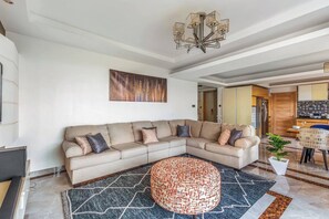A modern living room with a sectional sofa, a patterned ottoman, a chandelier, and a kitchen area in the background. - Urban Oasis (Nairobi)