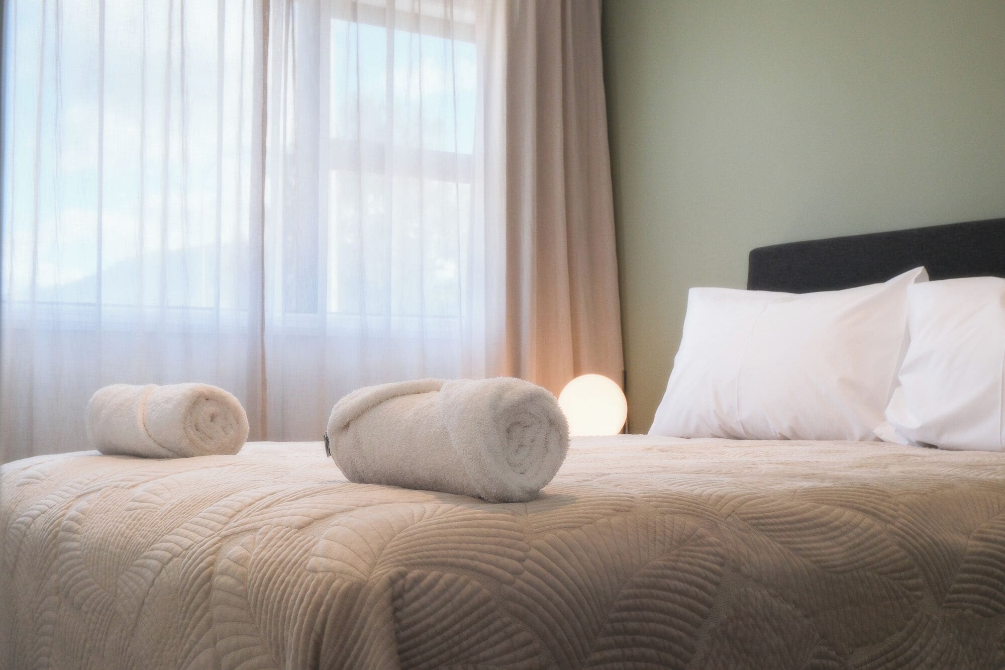 Une chambre d’hôtel avec un lit, deux serviettes enroulées, une tête de lit et une fenêtre avec des rideaux légers.