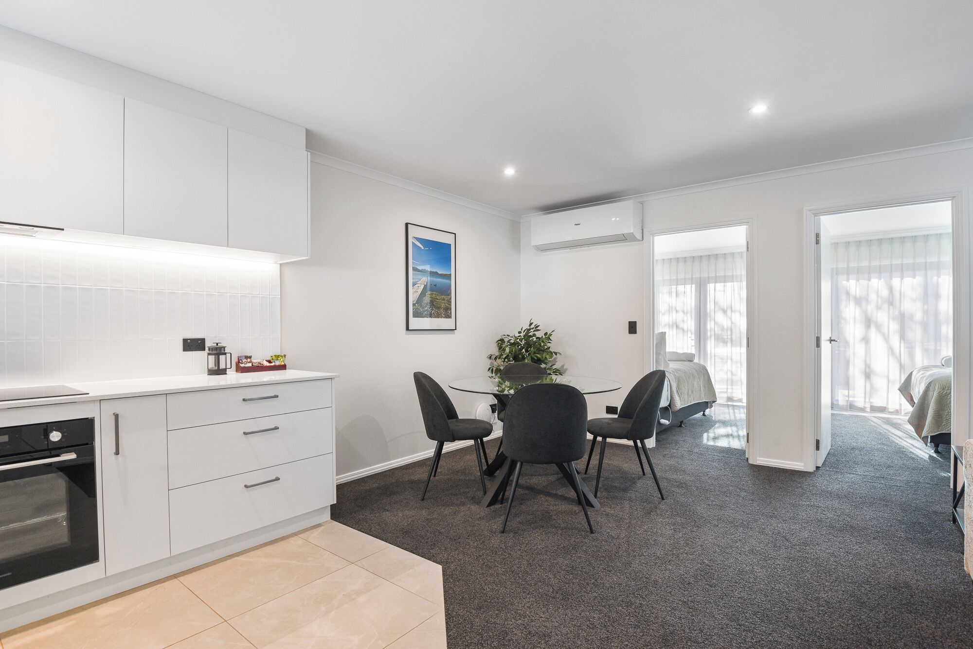 A modern kitchen with white cabinetry, a dining area with a glass table and chairs, and a bedroom with a large window.