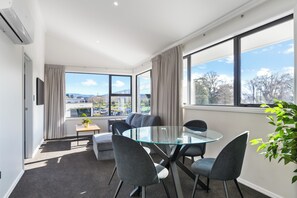 Habitación de hotel moderna con una mesa de vidrio, sillas grises, un pequeño sofá, televisión y vistas a un estacionamiento y árboles.