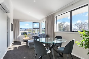 A modern hotel room with a glass table, grey chairs, a small sofa, a TV, and a view of a parking lot and trees.