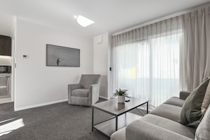 Una sala de estar moderna con un sofá gris, un sillón a juego, una mesa de centro y un amplio ventanal con cortinas traslúcidas.