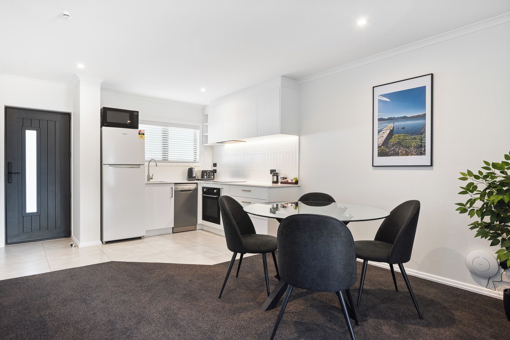 Een praktische keuken met moderne apparatuur, een eethoek met een glazen tafel en stoelen, en een lijst met een landschapsafbeelding aan de muur.