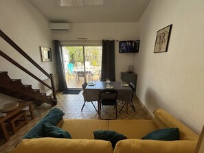 A living room with a yellow sofa, a staircase, a dining area with a table and chairs, and a TV on the wall.