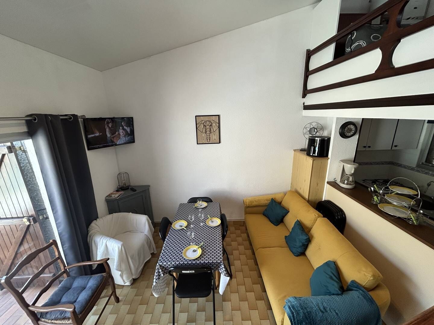 Un espace de vie compact comprenant une table à manger pour quatre personnes, un canapé, un téléviseur fixé au mur et un coin cuisine avec une cuisinière et des placards.