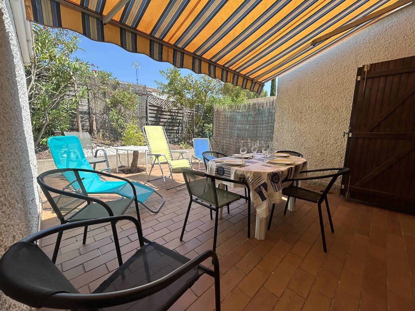Un patio exterior con una mesa lista para comer, rodeado de sillas y un área cubierta con un toldo a rayas.