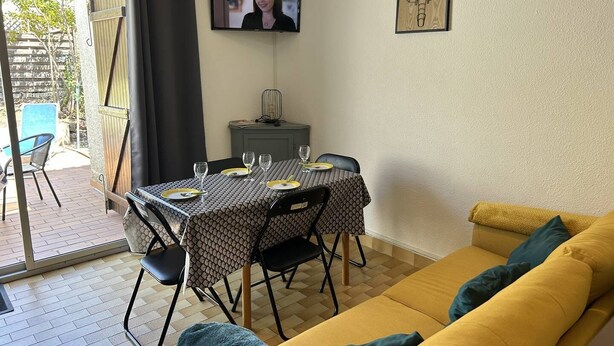 Una zona de menjador amb una taula preparada per a dues persones, un televisor fixat a la paret i un sofà amb coixins.