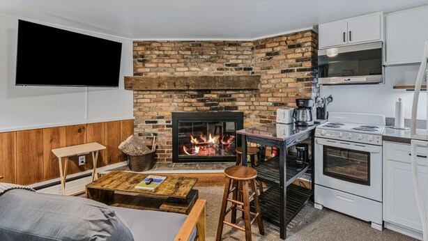 A cozy living area with a brick fireplace, a flat-screen TV mounted on the wall, a wooden coffee table, and a kitchen with white cabinets and appliances. - First Floor Retreat | Minutes to Stowe Mountain (Stowe)