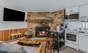 Een gezellige zithoek met een metselwerk openhaard, een flatscreen televisie aan de muur, een houten salontafel, en een keuken met witte kasten en apparatuur.