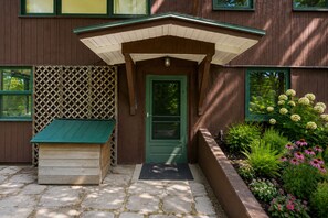 Ein Holzhaus mit grüner Tür, einer kleinen Veranda mit Bank und einem Garten mit verschiedenen Pflanzen und Blumen.