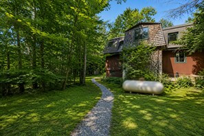 A house with a gravel path leading to a wooden door, surrounded by lush greenery and trees. - First Floor Retreat | Minutes to Stowe Mountain (Stowe)