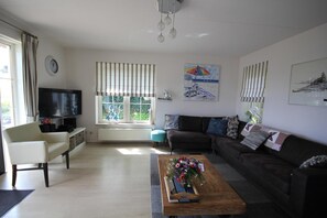 Ein Wohnzimmer mit einer Couch, einem Couchtisch mit Blumen, einem Fernseher und einem Fenster mit Vorhängen.