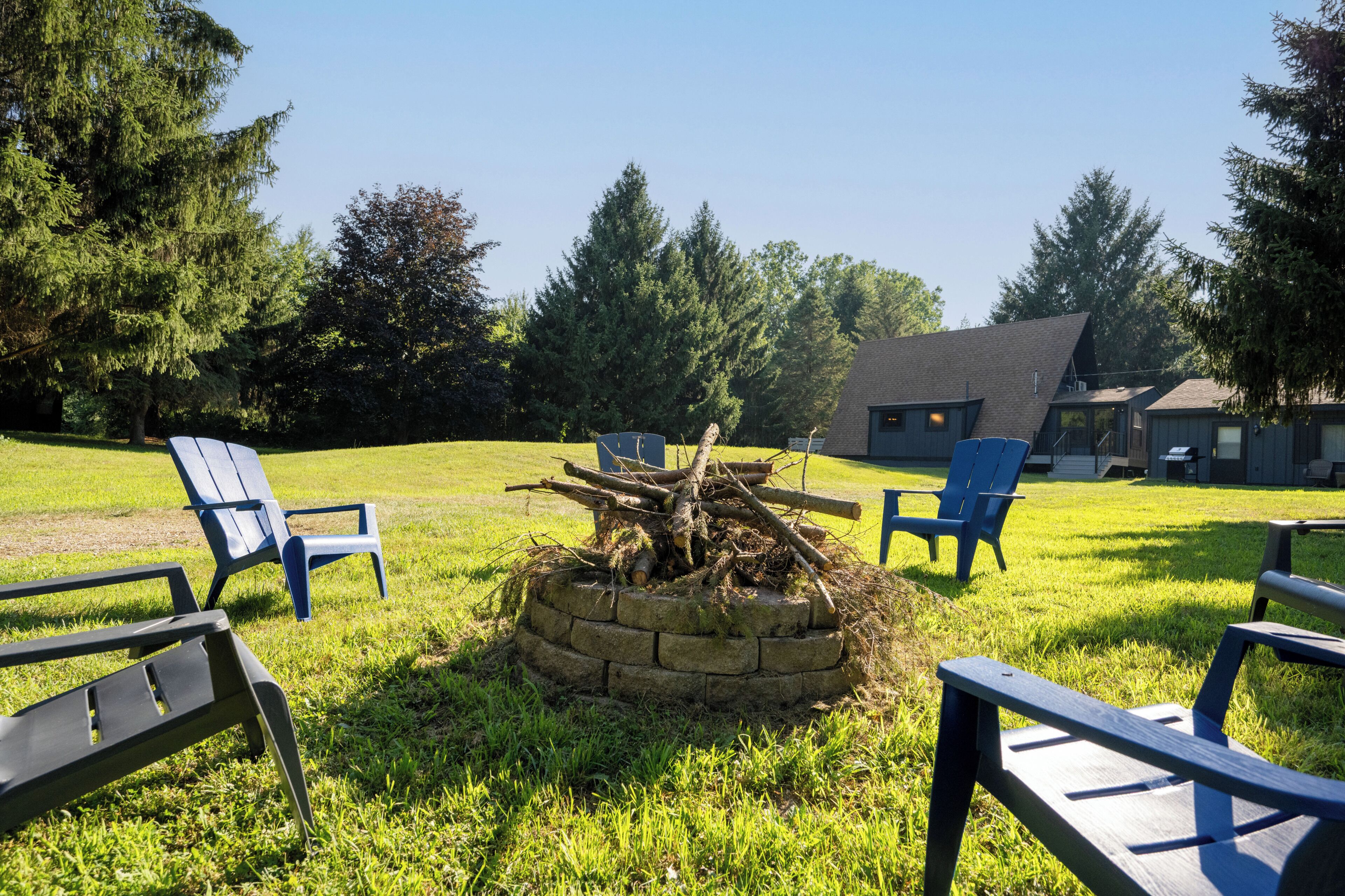 A grassy outdoor area with a stone fire pit, several blue chairs, and a building with a sloped roof in the background.