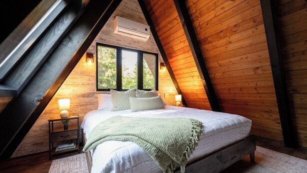 Ett sovrum med ett sluttande tak, en säng med kuddar och ett täcke, två sänglampor och ett fönster med utsikt över träd.