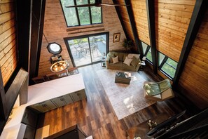A spacious living area with a dining table and chairs, a sofa, and a large window providing a view of the outdoors. - Secluded A-Frame on 10 Acres | At the Pines (Fennville)