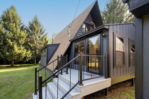 A modern house with a sloped roof, large windows, and a wooden deck with a metal railing. - Secluded A-Frame on 10 Acres | At the Pines (Fennville)