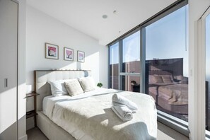 Ein modernes Schlafzimmer mit einem großen Bett, einem Nachttisch, einer Nachttruhe und einem großen Fenster mit Blick auf die Stadt.
