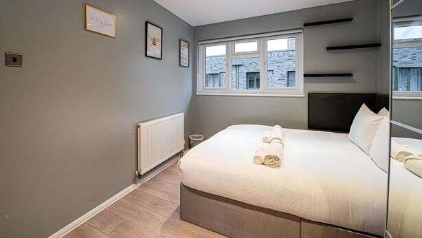 Une chambre moderne avec un grand lit, une fenêtre donnant sur des immeubles, un radiateur et une étagère fixée au mur.