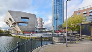 Un quartier riverain urbain moderne, avec un bâtiment remarquable caractérisé par une section en porte-à-faux, un gratte-ciel en verre imposant et une promenade piétonne bordée d’une clôture.