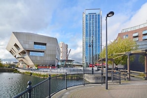 A modern urban waterfront with a distinctive building featuring a cantilevered section, a tall glass skyscraper, and a pedestrian walkway with a fence. - Beautiful flat (London)
