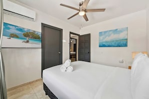 Una habitación de hotel con una cama, un ventilador de techo, un equipo de aire acondicionado, una puerta y dos cuadros en las paredes.