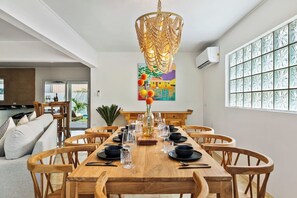Un comedor con una mesa de madera dispuesta para seis personas, rodeada de sillas de madera, una lámpara decorativa colgante y un cuadro colorido en la pared.
