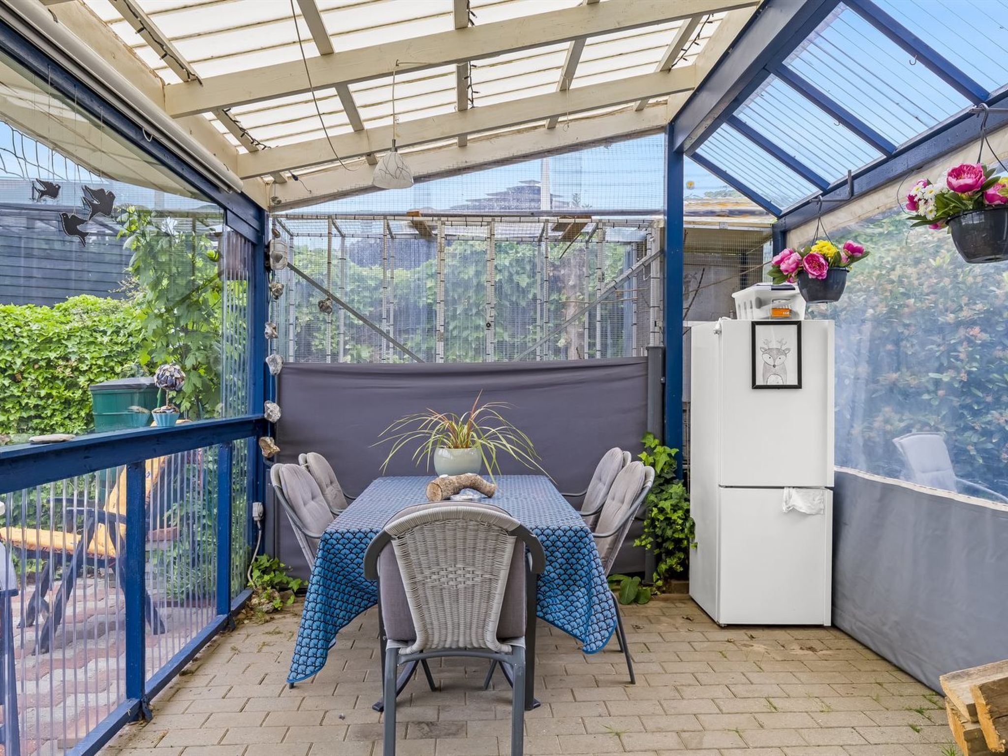 A covered patio with a table and chairs, a white refrigerator, and hanging flower baskets.