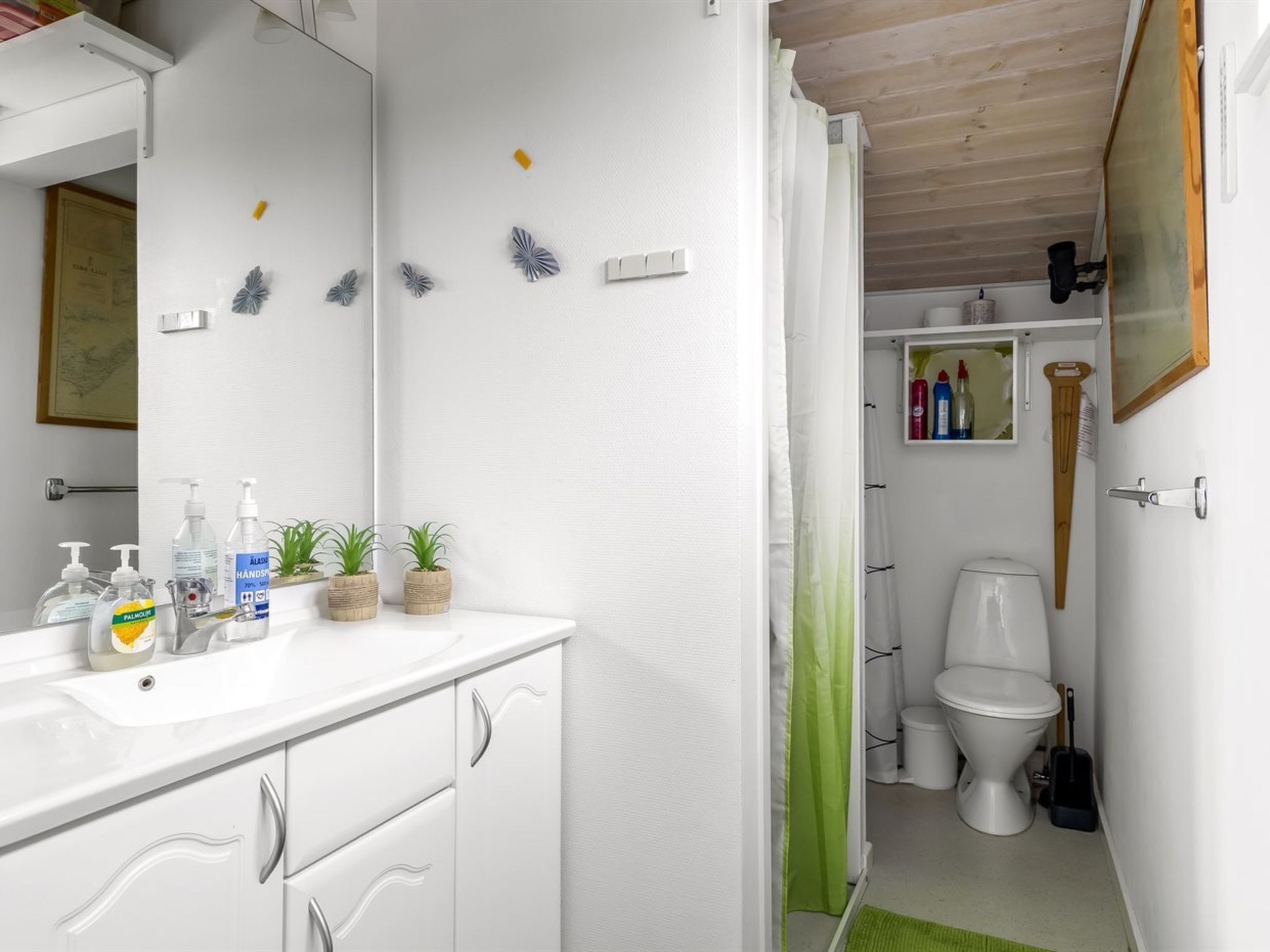 Casa de banho com lavatório branco, espelho e sanita. Há artigos de higiene pessoal no lavatório e uma cortina de chuveiro.