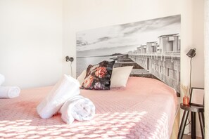 Un lit bien fait, agrémenté de serviettes blanches roulées, d’un coussin à motifs floraux et d’un grand tableau représentant un paysage côtier.